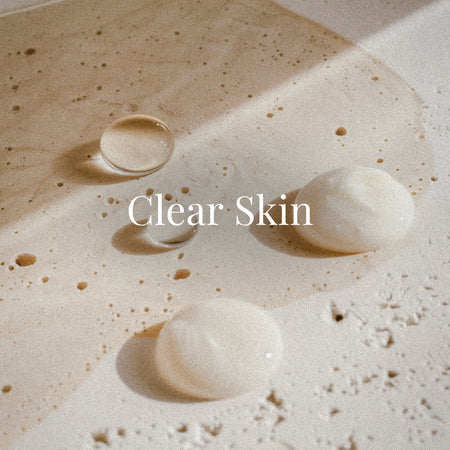 Creamy serum drops rest atop a smooth beige stone surface lightly splashed with water and product. The scene feels clean and minimal. White serif text centered on the image reads “Clear Skin.”